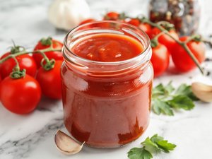 Im Vordergrund ein offenes Glas mit Tomatensoße. Dahinter frische Tomaten und Gewürze.