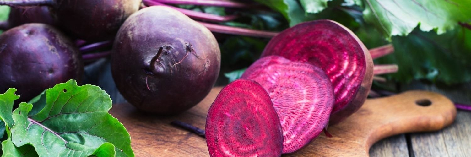 Auf einem Holzbrett liegen rote Beteknollen mit Blättern daran und eine Rote Bete ist in Scheiben geschnitten.
