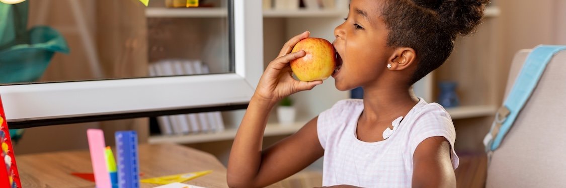 Eine Schülerin sitzt zuhause an ihrem Schreibtisch und beißt in einen Apfel.