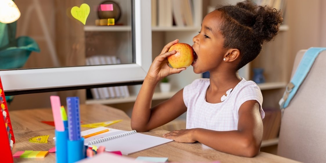 Eine Schülerin sitzt zuhause an ihrem Schreibtisch und beißt in einen Apfel.. Artikel "Was hat mein Apfel mit dem Klima zu tun?" öffnen.