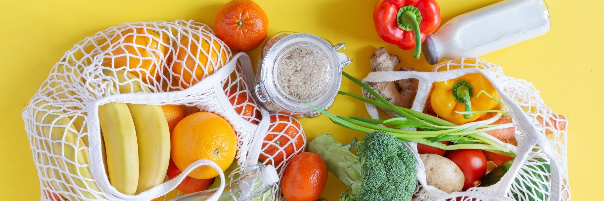 Zwei Einkausnetze auf gelben Hintergrund, gefüllt mit Obst, Gemüse und Reis, Nudeln sowie Milch in Glasgefäßen.