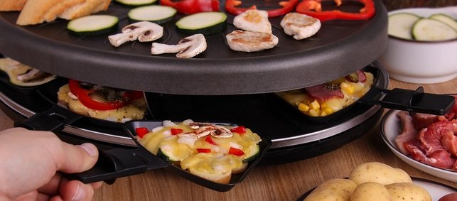 Auf einem Raclette-Grill werden Champignonscheiben, Zucchinischeiben. Paprikaringe und Fleisch gegrillt. Eine Hand nimmt ein Pfännchen aus dem Grill. Darin befinden sich mit Käse überbackene Kartoffeln mit Paprika, Zwiebeln und Champignons. Vor dem Grill stehen Schalen mit Kartoffeln, Fleisch und Zucchini.   