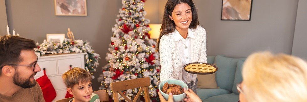 Eine Familie feiert Weihnachten zuhause. Im Hintergrund steht ein Weihnachtsbaum und der Tisch, an dem ein Mann, ein Junge und eine ältere Frau sitzen, ist festlich geschmückt. Eine Frau bringt Weihnachtsessen zum Tisch. 