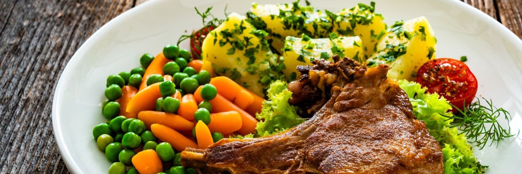 Auf einem weißen Teller ist ein gegrilltes Schweinekotelett mit Gemüse und Kartoffeln angerichtet.