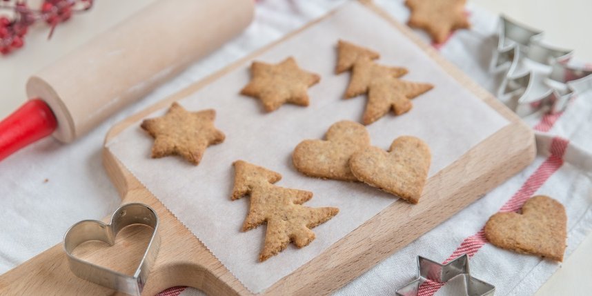 Auf einem Holzbrett liegen auf Backpapier ausgestochene Plätzchen mit Weihnachtsmotiven, daneben liegen Ausstechformen und ein Nudelholz. . Artikel "Weihnachten in der Küche: Einfach besser backen " öffnen.