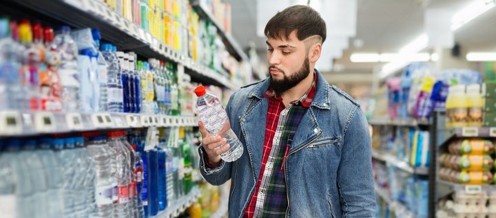 Ein junger Mann steht im Supermarkt vor dem Getränkeregal. Er hält eine Wasserflasche in der Hand und liest das Etikett. 