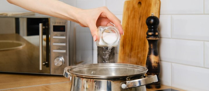 Eine Frauenhand schüttet Salz aus einem Glasgefäß in einen Topf auf dem Herd. Im Hintergrund ist eine Pfeffermühle, ein Holzbrett und eine Mikrowelle.