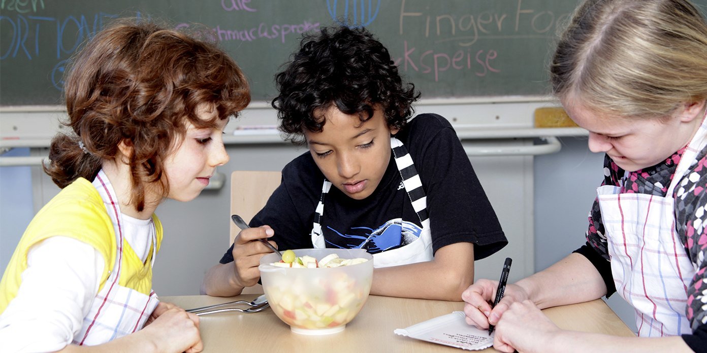 Drei Kinder schneiden im Klassenzimmer Obst.. Artikel "Ernährungsbildung" öffnen.