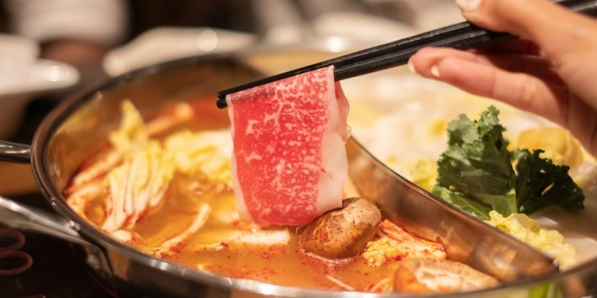 Ein Metalltopf gefüllt mit Brühe, Pilzen, Gemüse und Fleisch. Eine Hand hält an zwei Stäbchen eine dünne Scheibe Fleisch darüber.. Artikel "„Hot Pot“ aus Asien" öffnen.