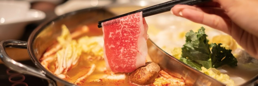 Ein Metalltopf gefüllt mit Brühe, Pilzen, Gemüse und Fleisch. Eine Hand hält an zwei Stäbchen eine dünne Scheibe Fleisch darüber.