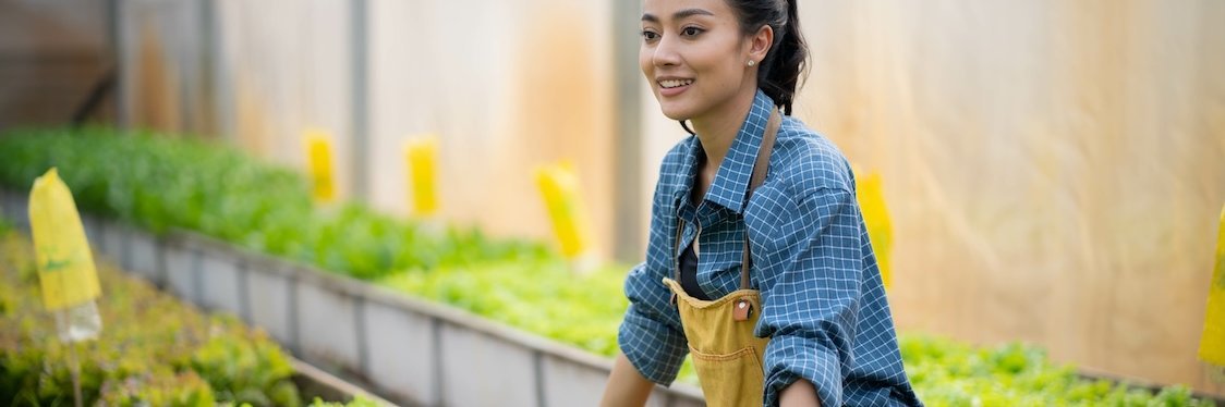 Eine junge Frau in Arbeitskleidung lehnt lachend an einem Pflanzbeet.