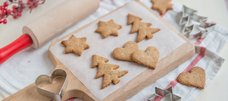 Auf einem Holzbrett liegen auf Backpapier ausgestochene Plätzchen mit Weihnachtsmotiven, daneben liegen Ausstechformen und ein Nudelholz. 