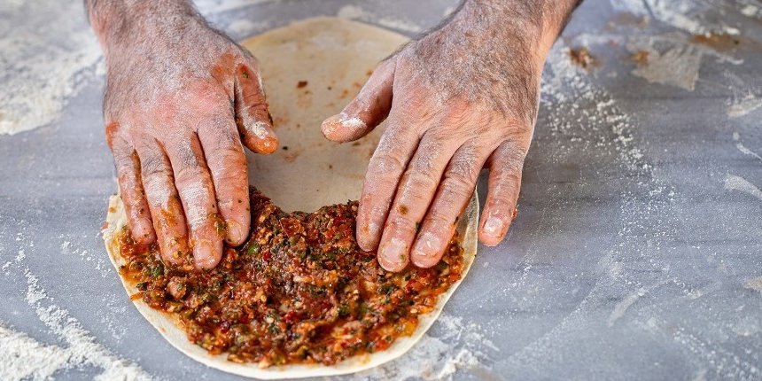 Auf einer bemehlten Arbeitsfläche liegt ein Lahmacun-Fladen. Zwei Hände verteilen darauf den Belag. . Artikel "Lahmacun selbst gemacht" öffnen.