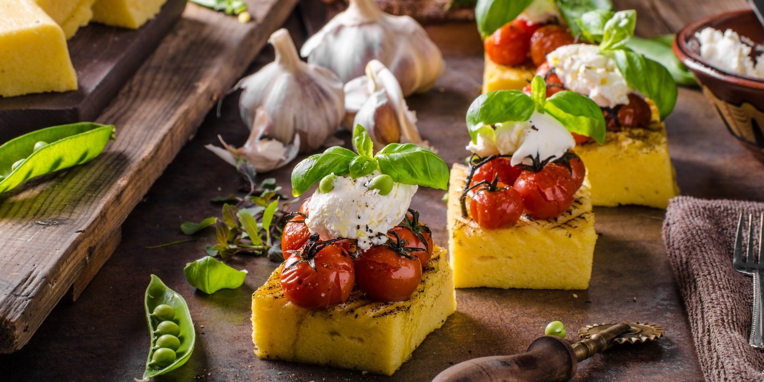 Auf einem dunklen Untergrund liegen gegrillte Polentastücke mit gebackenen Kirschtomaten, Mozzarella, Basilikum und Erbsen angerichtet. . Artikel "Polenta – das Maisgericht aus Italien" öffnen.