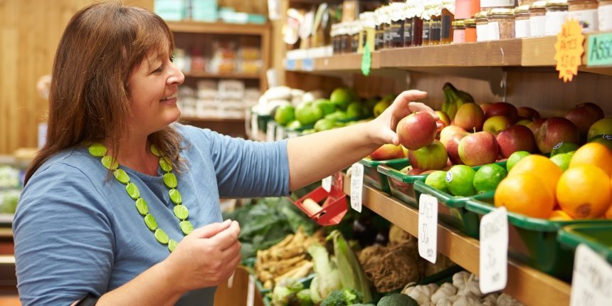 Eine Frau steht vor einem Regal im Hofladen und greift nach einem Apfel.. Artikel "Wie gelingt der bewusste Einkauf?" öffnen.