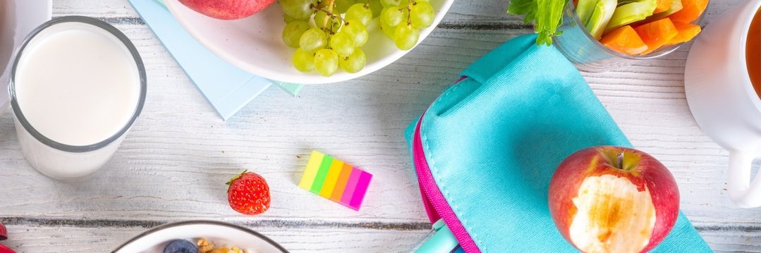 Auf einem Tisch stehen ein Müsli, Äpfel, Rohkost, ein Glas Milch, eine Federmappe und Stifte.