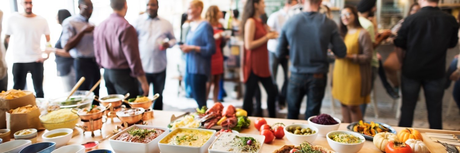 Im Vordergrund ist ein buntes, vegetarisches Buffet zu sehen, im Hintergrund stehen Menschen und unterhalten sich.