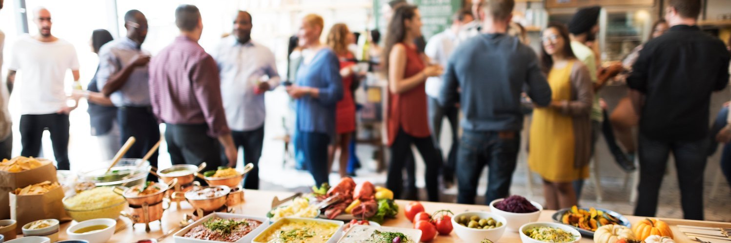 Im Vordergrund ist ein buntes, vegetarisches Buffet zu sehen, im Hintergrund unterhalten sich Menschen.