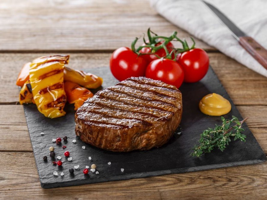 Ein gegrilltes Steak auf einer Schieferplatte, angerichtet mit gegrillter Paprika, Strauchtomaten, Senf, Thymian, Pfeffer und Salz.