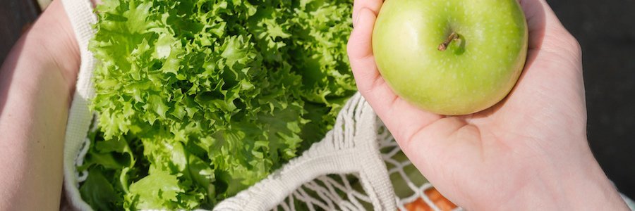 Eine Hand greift in einen Stoffbeutel, in dem sich ein Apfel, ein Salatkopf und anderes Gemüse befindet.