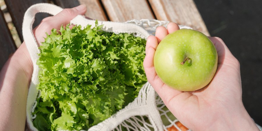 Eine Hand greift in einen Stoffbeutel, in dem sich ein Apfel, ein Salatkopf und anderes Gemüse befindet.. Artikel "Was hat unser Essen mit dem Klima zu tun?" öffnen.