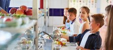 Mehrere Schulkinder stehen mit Tabletts an der Essensausgabe an und eins bekommt Essen mit einer großen Kelle auf seinen Teller geschöpft. 