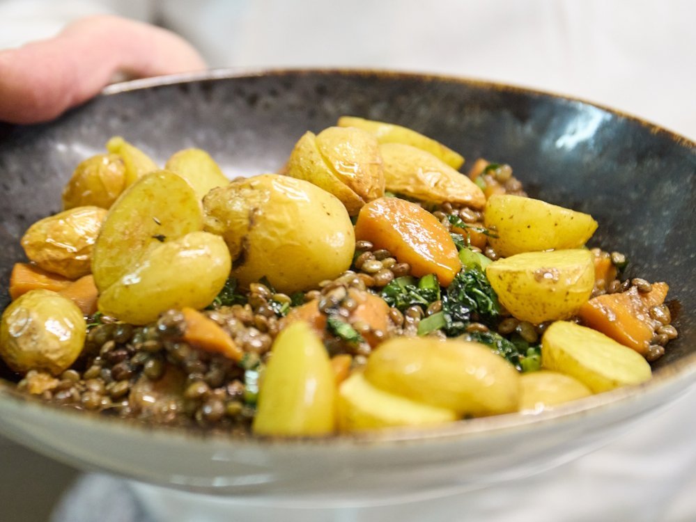 Ein schwarzer Teller mit einem Gericht aus Kartoffeln, Moehren und Linsen.
