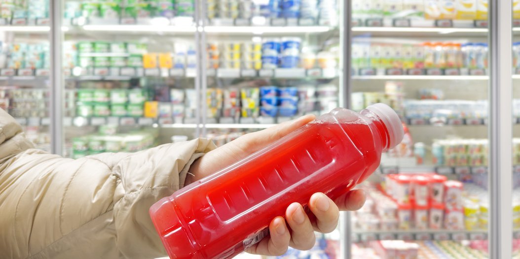 Eine Hand hält ein rotes Getränk. Im Hintergrund ist ein Kühlregal in einem Supermarkt zu sehen.. Artikel "Dem Zucker auf der Spur" öffnen.