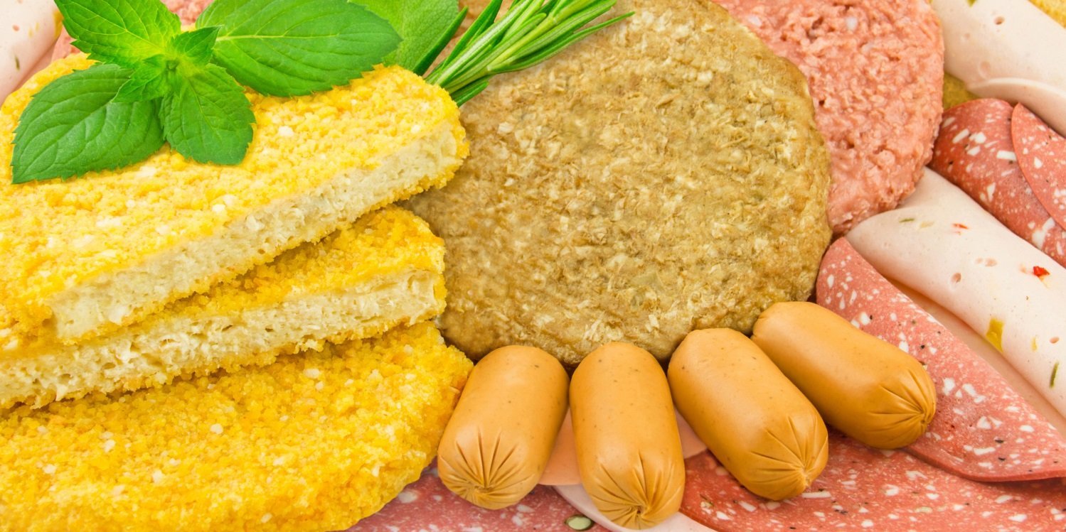 Eine Fläche ist bedeckt mit verschiedenen pflanzliche Patties, Aufschnittsorten und Aufstrich in Wurstform, dekoriert mit Basilikumblättern und Rosmarin.. Artikel "Sind Alternativprodukte gut für Klima und Umwelt?" öffnen.
