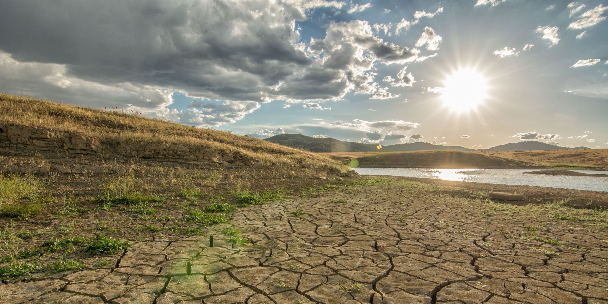 Eine ausgetrocknete Landschaft. Im Hintergrund ein See mit niedrigem Wasserstand. Und darüber scheint eine gleißende Sonne.. Artikel "Klima und Wandel - was jetzt zu tun ist" öffnen.