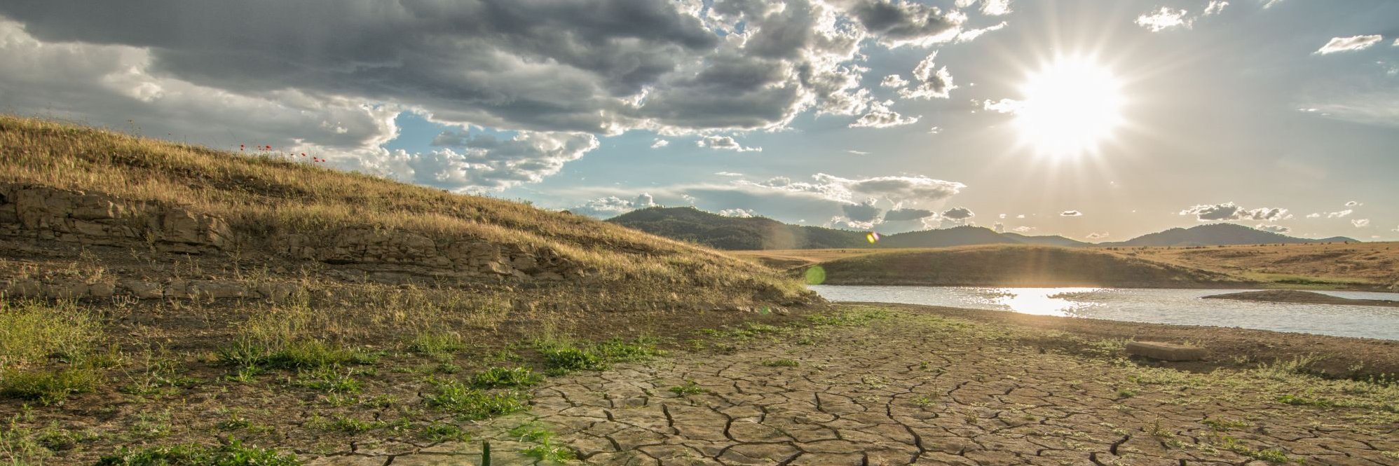 Eine ausgetrocknete Landschaft. Im Hintergrund ein See mit niedrigem Wasserstand. Und darüber scheint eine gleißende Sonne.