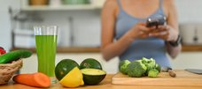 Eine Frau steht in der Küche vor der Arbeitsplatte. Auf dieser sind ein großes Glas mit einem grünen Getränk, eine Gurke, Möhre, Zitrone, Avocados und Brokkoli zu sehen. 