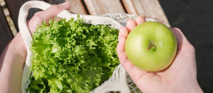 Hände halten klimafreundlichen Einkaufsbeutel mit Apfel und Salat.