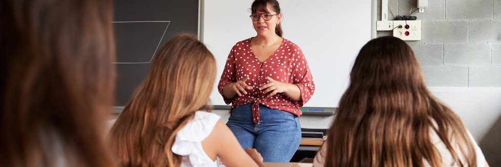 Eine Frau mittleren Alters mit Zopf und gepunkteter Bluse und Jeans sitzt auf einem Tisch. Im Vordergrund sitzen drei Kinder mit langen Haaren, man sieht sie von hinten. Im Hintergrund eine Tafel und ein Whiteboard.