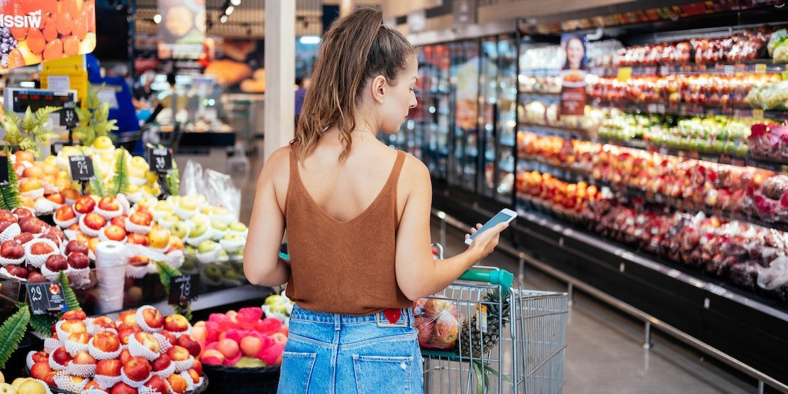 Frau steht vor einem Obst- und Gemüseregal und schaut auf ihr Telefon.. Artikel "Verkaufstricks im Supermarkt" öffnen.