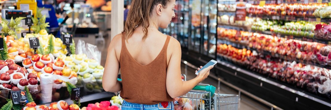 Frau steht vor einem Obst- und Gemüseregal und schaut auf ihr Telefon.