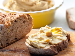 Eine Scheibe Brot mit Hummus. Daneben ein Brot und dahinter eine Schüssel mit Hummus.