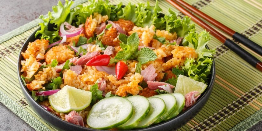 Auf einer Bastmatte steht ein dunkler Teller mit Salatblättern, knusprigem Reis, Gurkenscheiben, Limettenstücken, Chilischoten und Minzeblättern. Daneben liegt ein Paar Essstäbchen.. Artikel "„Crispy Rice Salad“" öffnen.