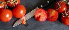 Rote Tomaten liegen auf schwarzer Schieferplatte. Eine ist halbiert. Auf einer Messerklinge, die zwischen den Tomaten hervorlugt, liegt der herausgeschnittene Stielansatz der halbierten Tomate.
