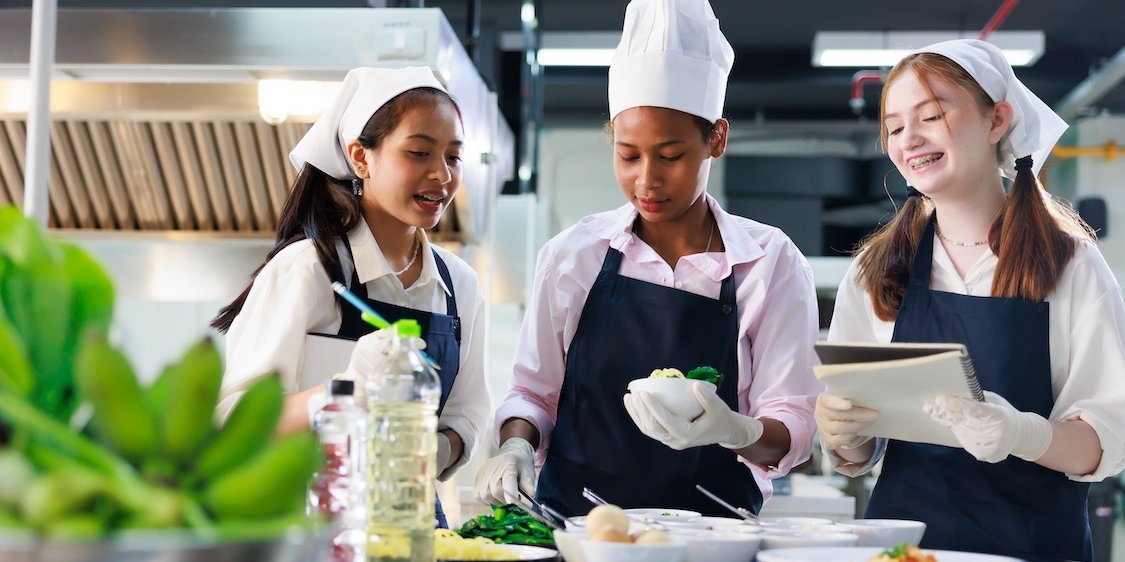 Drei Schülerinnen mit Kochmützen und Schürzen kochen in einer professionellen Küche.. Artikel "Schulessen ist Teil der Ernährungsbildung" öffnen.