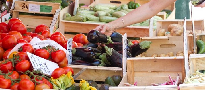 Eine Frau kauft Gemüse auf dem Markt. In Holzkisten liegen Tomaten, Auberginen, Zucchini und Kartoffeln vor ihr. 