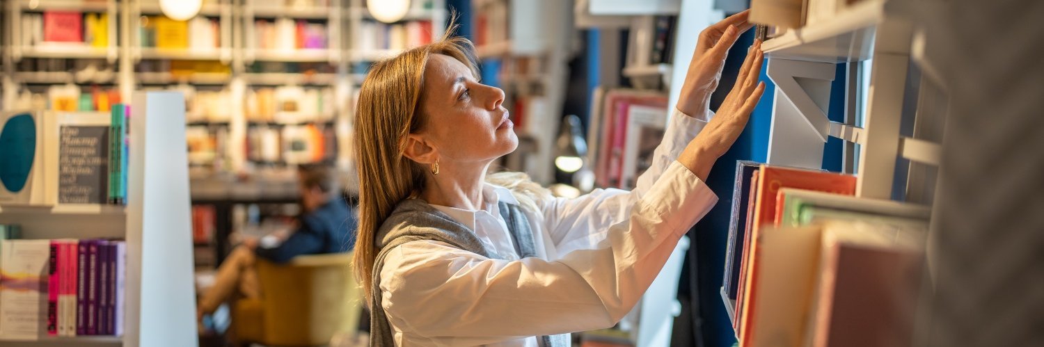Eine Frau steht vor einem Bücherregal und sucht sich ein Buch aus.