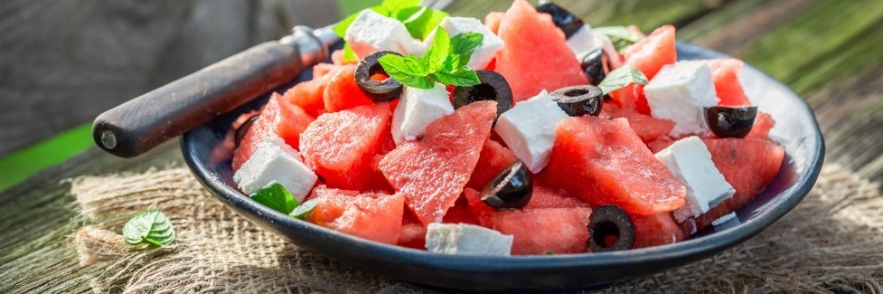 Auf einem Holztisch im Freien steht ein dunkler Teller mit gewürfelter Wassermelone, Fetastücken, schwarzen Oliven und Minzblättern. 