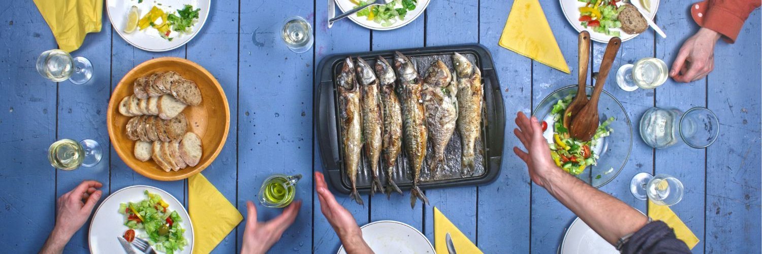Auf einem blauen Holztisch liegt eine Platte mit gegrilltem Fisch, eine Salatschüssel und ein Korb mit Brot. Drumherum stehe weiße Teller und Gläser und Menschen greifen nach dem Essen.