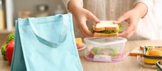 Auf einer Arbeitsfläche in der Küche steht eine hellblaue Tasche. Im Hintergrund sieht man Hände, die einen Sandwich belegt mit Salat in eine Dose legen. 