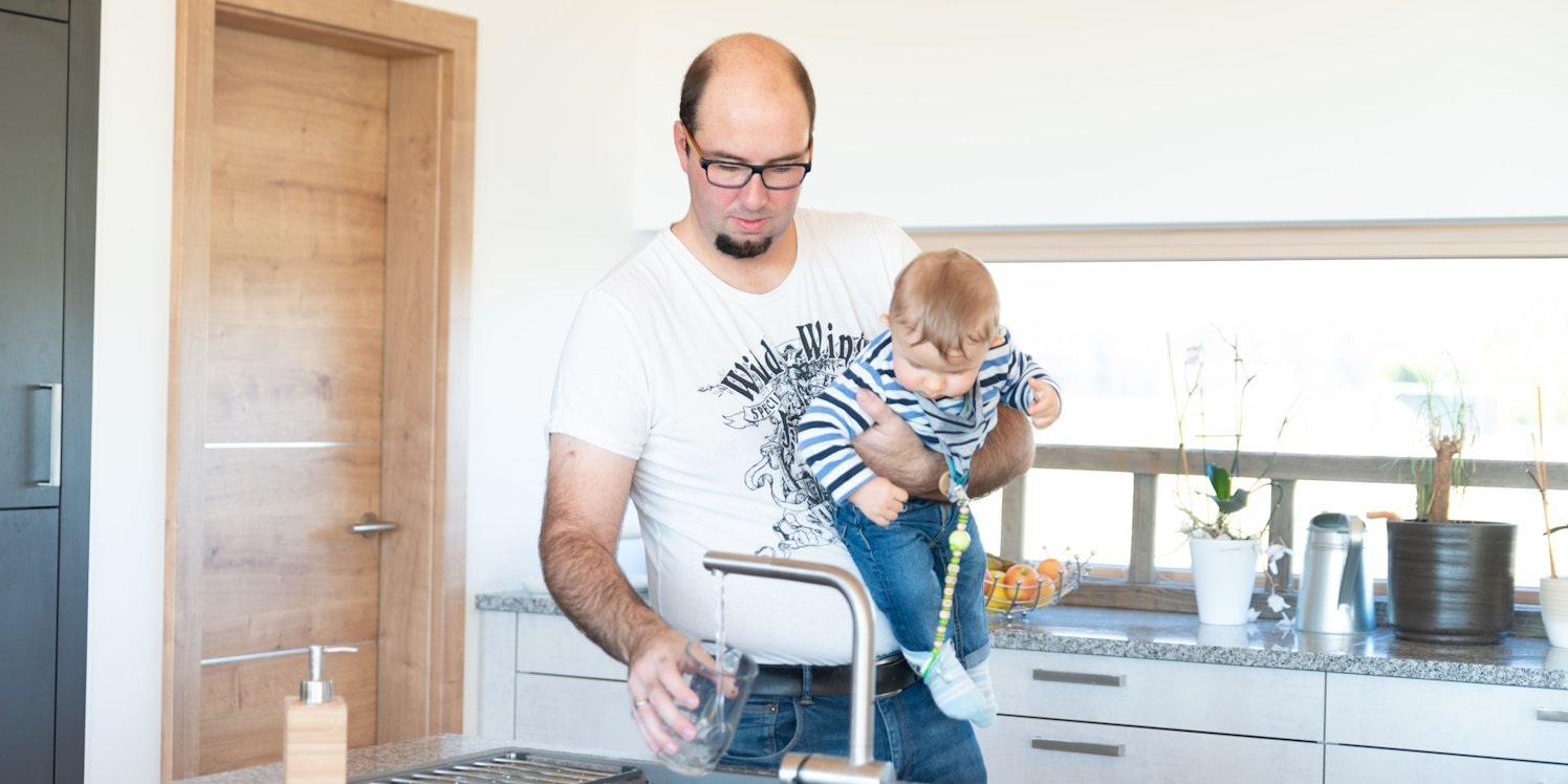 Ein Mann hat einen Baby auf dem Arm und lässt Wasser aus einem Hahn in ein Glas laufen.. Artikel "Wasser: Gesundheit und Umwelt" öffnen.