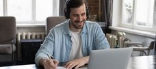 Ein bärtiger Mann mit Headset sitzt vor einem Laptop und schaut lächelnd auf den Bildschirm während er etwas auf einem Blatt notiert.
