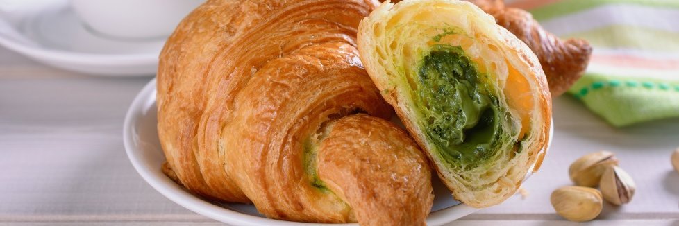 Auf einem Teller liegt ein aufgeschnittenes Croissant mit Pistaziencreme gefüllt. Daneben liegen einige Pistazien und eine gestreifte Serviette neben einer Kaffeetasse.