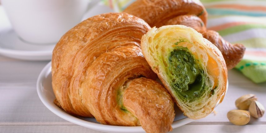 Auf einem Teller liegt ein aufgeschnittenes Croissant mit Pistaziencreme gefüllt. Daneben liegen einige Pistazien und eine gestreifte Serviette neben einer Kaffeetasse.. Artikel "Süßer Sommerhype um die Pistazie" öffnen.