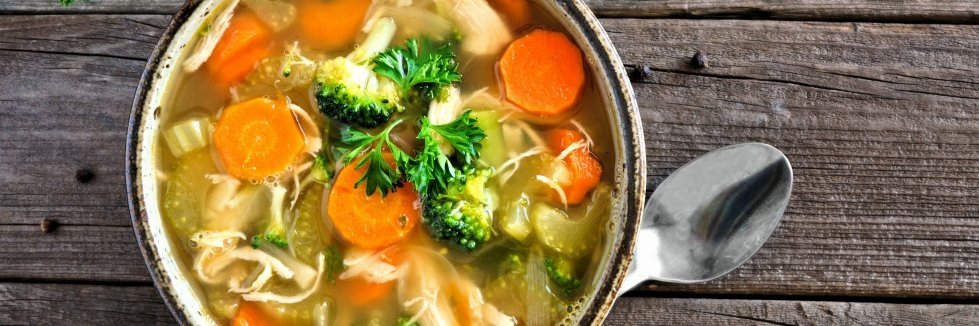 Auf einem dunklen Holztisch steht eine weiße Suppenschale mit Hühnersuppe, Möhren, Stangensellerie, Brokkoli und Petersilie. Daneben liegt ein Löffel.